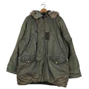 Op de afbeelding: Een olijfgroene parka met een met bont gevoerde capuchon. De jas heeft meerdere zakken, knoopsluitingen en een patchdetail op de borst. De parka hangt aan een houten hanger.