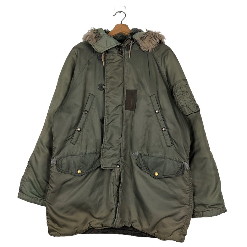 Op de afbeelding: Een olijfgroene parka met een met bont gevoerde capuchon. De jas heeft meerdere zakken, knoopsluitingen en een patchdetail op de borst. De parka hangt aan een houten hanger.