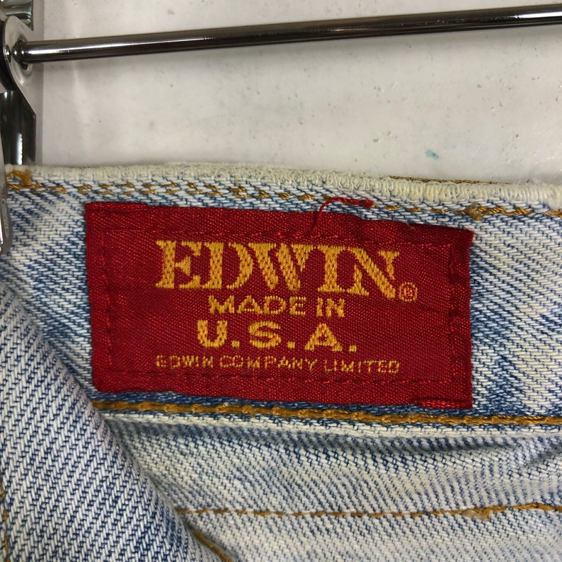 Może przedstawiać: Czerwona metka z białym tekstem, na kt&oacute;rej widnieje napis "EDWIN MADE IN U.S.A. EDWIN COMPANY LIMITED" na parze jasnoniebieskich dżins&oacute;w.