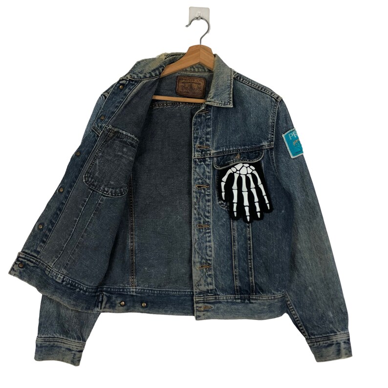 Puede incluir: Una chaqueta vaquera azul con un parche de mano de esqueleto en el bolsillo izquierdo y un parche azul con texto blanco en el bolsillo derecho. La chaqueta tiene cierre de botones y mangas largas.