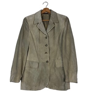 Könnte beinhalten: Ein hellbrauner Wildlederblazer mit klassischem Reverskragen und Dreiknopfverschluss. Die Jacke hat zwei Vordertaschen und hängt an einem Holzbügel. Der Blazer hat eine taillierte Passform.