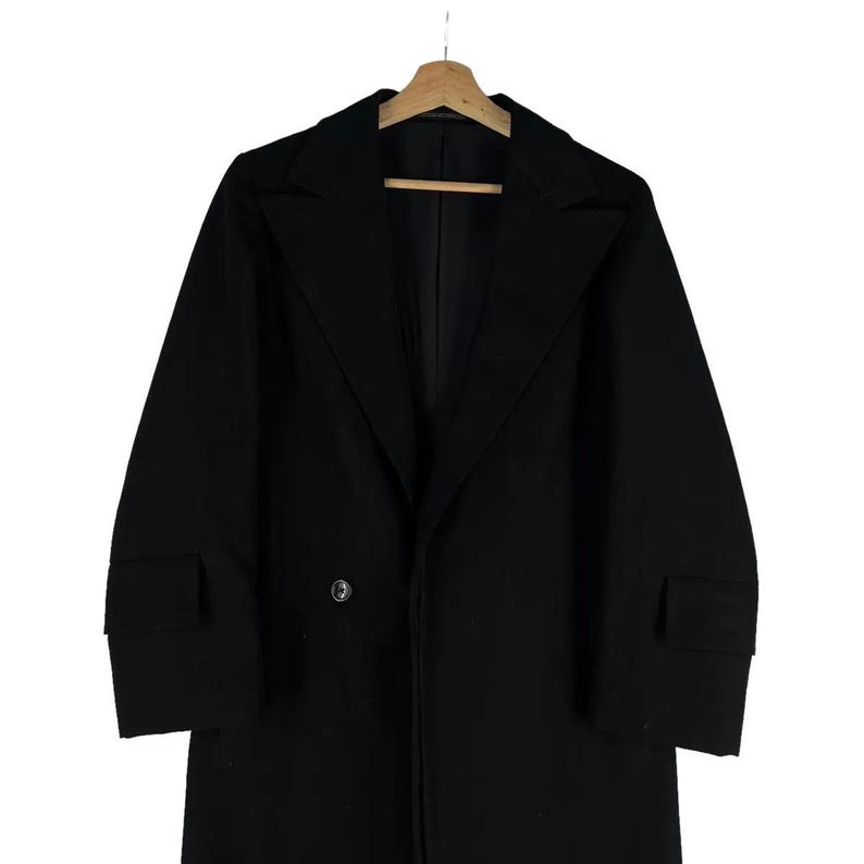 May include: A black overcoat with a notched collar and two front pockets. The coat has a double-breasted closure with two buttons and is hanging on a wooden hanger.