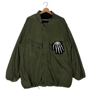 May include: A green reversible jacket with a skeleton hand patch on the front pocket. The jacket has a button-up front and a drawstring at the bottom.