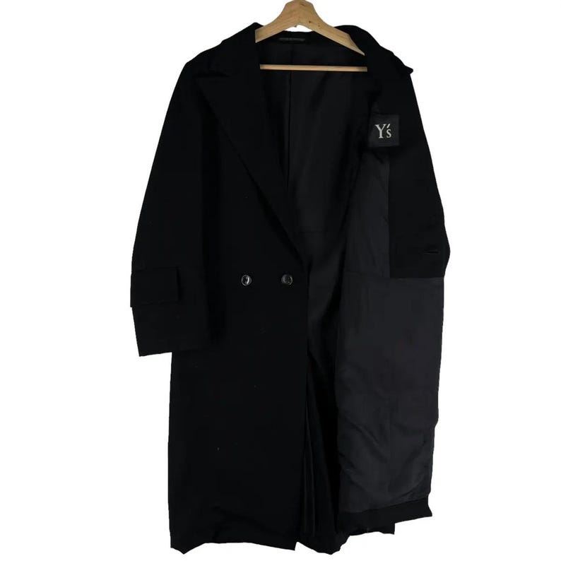 May include: A long, black double-breasted coat with a notched lapel and a partial open design. The coat is hanging on a wooden hanger. A small white tag with the text "Y's" is visible on the coat.