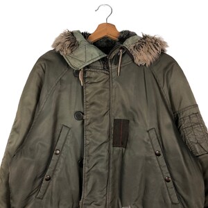 Op de afbeelding: Olijfgroene winterjas met een met imitatiebont gevoerde capuchon. De jas heeft een knoopsluiting, een ritssluiting en meerdere zakken. De jas hangt aan een houten kleerhanger.