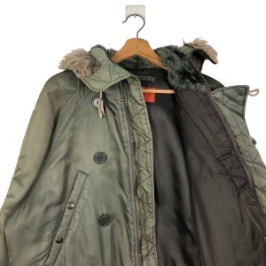 Op de afbeelding: Een olijfgroene winterjas met een met bont gevoerde capuchon en een dubbele rij knopen. De jas hangt aan een houten kleerhanger. De binnenvoering is donkerbruin.