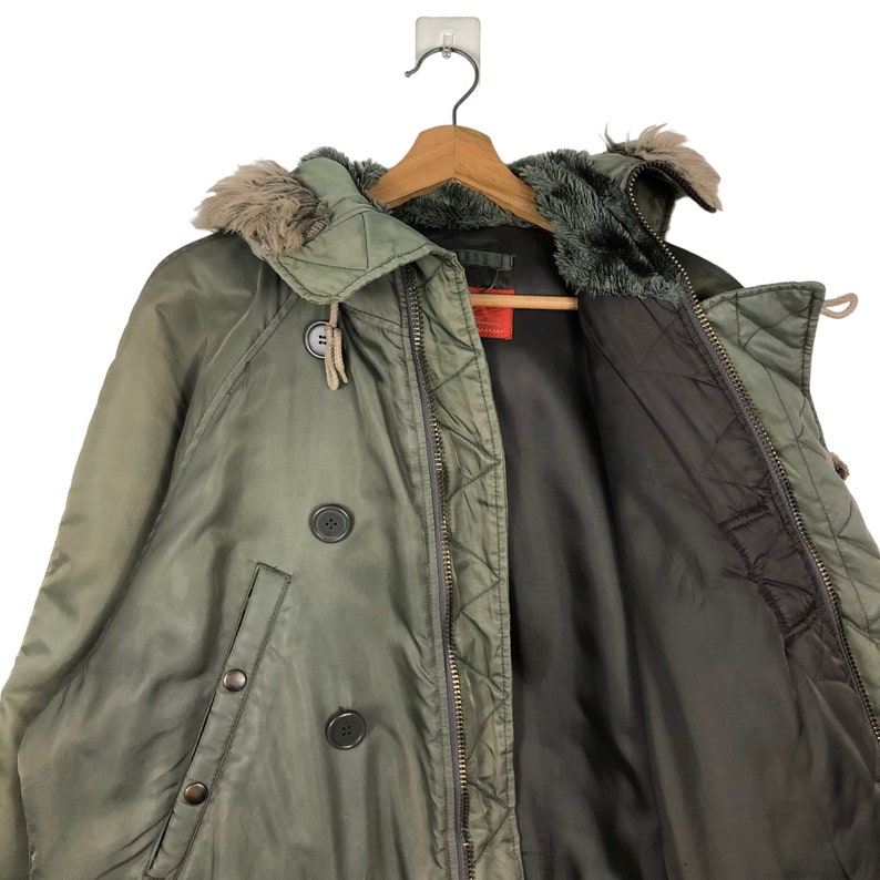 Op de afbeelding: Een olijfgroene winterjas met een met bont gevoerde capuchon en een dubbele rij knopen. De jas hangt aan een houten kleerhanger. De binnenvoering is donkerbruin.