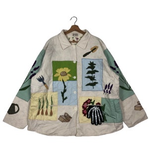Puede incluir: Chaqueta acolchada color crema con parches bordados con temática de jardín. Presenta un girasol, lavanda, herramientas de jardinería y una mano de esqueleto. La chaqueta tiene una parte delantera abotonada y mangas largas.