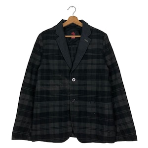 Puede incluir: Un blazer a cuadros gris oscuro y negro con un cuello y solapa grises en contraste. El blazer tiene dos botones y un bolsillo. La chaqueta está colgada de una percha de madera.