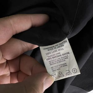 May include: A close-up of a tag with text in English and French, held by a hand. The tag reads "FABRIQUE AU JAPON MADE IN JAPAN 100% LAINE WOOL DOUBLURE LINING 100% RAYONNE RAYON". It also includes care instructions.