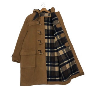 Puede incluir: Un abrigo tipo duffle de color marrón claro con forro a cuadros. El abrigo tiene cierres de madera, una capucha y dos bolsillos delanteros. Cuelga de una percha de madera.