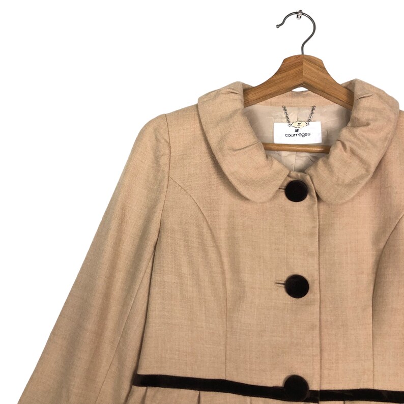 Op de afbeelding: Een beige jas met een ronde kraag en drie donkerbruine knopen. De jas heeft een donkerbruine fluwelen bies en een label met de tekst "courreges". Hij hangt aan een houten kleerhanger.