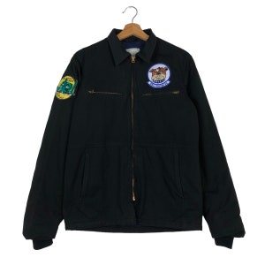 May include: A black work jacket with a zipper closure and two patch pockets. The jacket has two embroidered patches, one with a green airplane and the other with a cartoon bulldog.