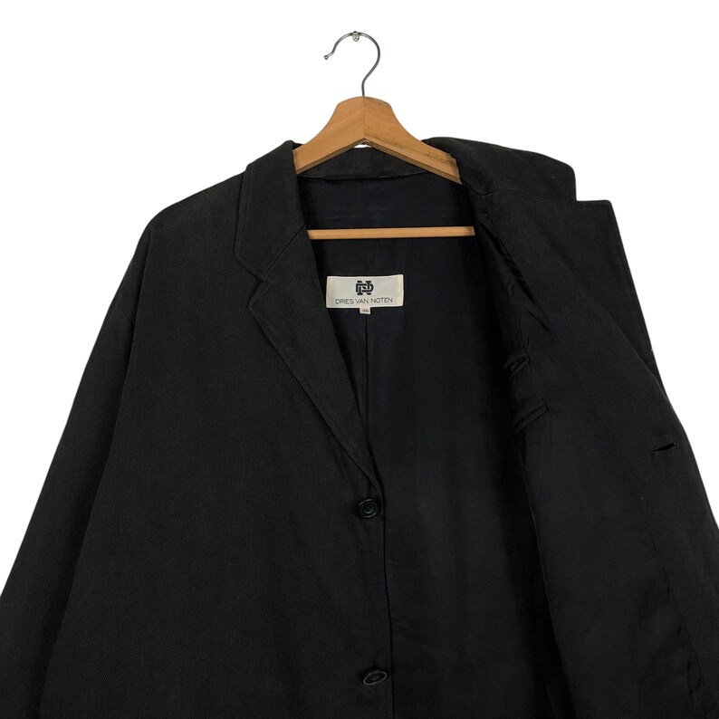 Może przedstawiać: Czarny blazer z pojedynczym zapięciem na guzik i metką z napisem "DRIES VAN NOTEN" wewnątrz.