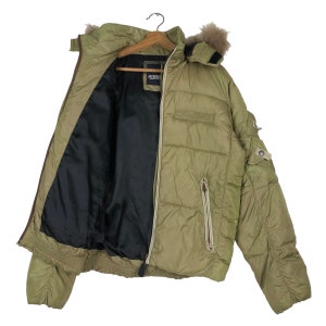 Puede incluir: Una chaqueta acolchada verde con capucha forrada de piel. La chaqueta tiene cierre de cremallera y un bolsillo con cremallera en el lado izquierdo. La chaqueta est&aacute; abierta y el forro es negro.