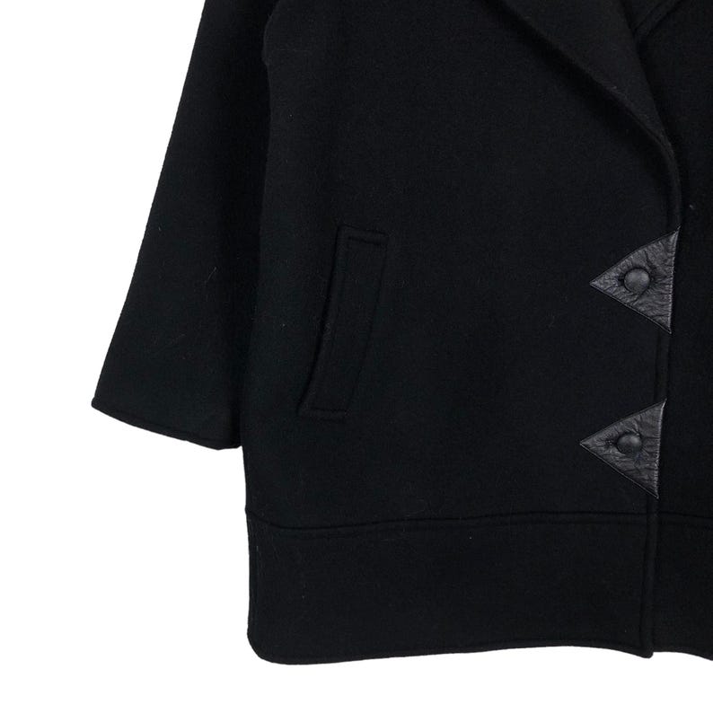 May include: Black wool coat with a notched collar, a single pocket, and three-quarter sleeves. The coat features leather triangle-shaped button closures. The hemline is straight and the coat appears to be a mid-length style.
