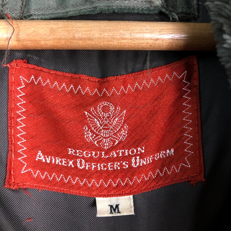 Op de afbeelding: Close-up van een rood Avirex Officer's Uniform label met witte tekst en een Amerikaans embleem. Het label is gestikt met witte draad en heeft maat "M" gedrukt op een klein wit label.