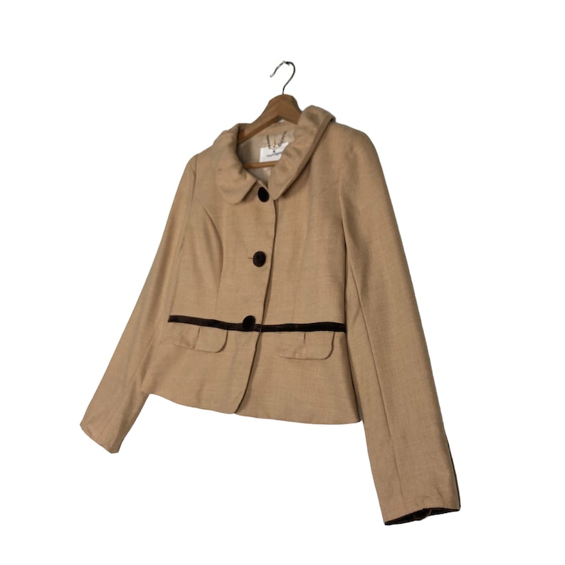 Op de afbeelding: Een beige blazer met een ronde kraag, zwarte knopen en twee voorzakken. De blazer heeft een donkerbruine bies langs de zakken en de kraag. De jas hangt aan een houten kleerhanger.