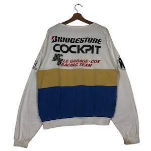 Pode incluir: Camisola de manga comprida branca, amarela, azul e vermelha com o texto "BRIDGESTONE COCKPIT LE GARAGE COX RACING TEAM" impresso nas costas.