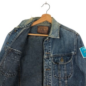 Puede incluir: Una chaqueta vaquera azul con un parche de cuero marr&oacute;n que dice "Edwin Co. Ltd." y un parche azul con el texto "Premium" en la manga derecha.