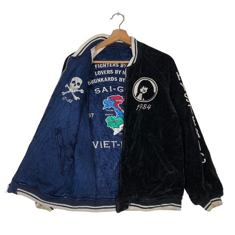 May include: A black and blue reversible bomber jacket with a skull and crossbones on one side and a woman's face with the text "1984" on the other side. The jacket also features the text "FIGHTERS BY LOVERS BY DRUNKARDS BY SAI-G VIET-H" and a map of Southeast Asia.