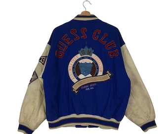 May include: Blue and cream varsity jacket with the text "GUESS CLUB" in red. The jacket features a large emblem on the back with the words "ALL WEATHER COLLECTION" and "CLASSIC SPORTS FOR ALL". The sleeves are cream-colored.