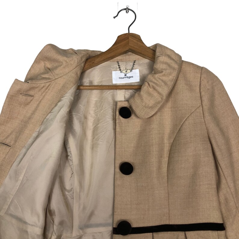 Op de afbeelding: Een beige jas met een ronde kraag en drie grote zwarte knopen. De jas heeft een zwarte bies en hangt aan een houten hanger. De binnenvoering is lichtbeige. Het label luidt "courreges".