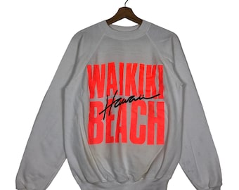 Op de afbeelding: Witte sweatshirt met de woorden "WAIKIKI BEACH Hawaii" in feloranje blokletters. Het woord "Hawaii" is in een cursief lettertype. De sweatshirt hangt aan een houten hanger.
