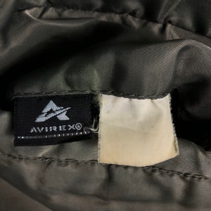 Op de afbeelding: Close-up van een donkergrijze stof met een zwart label met het Avirex-logo en tekst. Een blanco wit label is aan de stof bevestigd. De stof heeft een gewatteerde uitstraling.