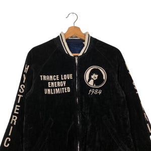 May include: Black velvet bomber jacket with white embroidered lettering on the front and sleeves. The lettering reads "TRANCE LOVE ENERGY UNLIMITED" and "HYSTERIC" on the sleeves. The jacket also features a white embroidered image of a woman with the text "1984" below it.