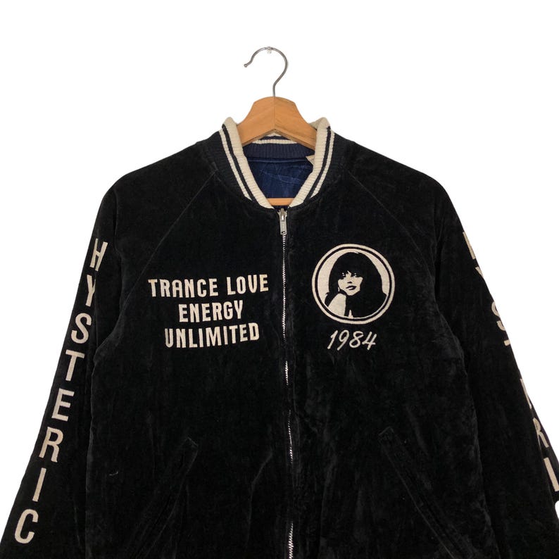 May include: Black velvet bomber jacket with white embroidered lettering on the front and sleeves. The lettering reads "TRANCE LOVE ENERGY UNLIMITED" and "HYSTERIC" on the sleeves. The jacket also features a white embroidered image of a woman with the text "1984" below it.