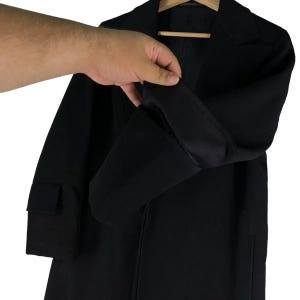 May include: A black coat with a notched lapel and long sleeves. The coat is made of a solid black fabric and has a pocket on the left side. The sleeve is being held up to show the lining.