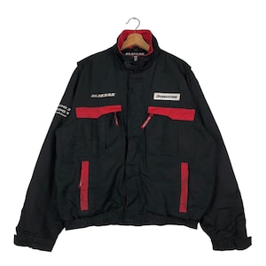 Könnte beinhalten: Schwarze Jacke mit roten Akzenten und den Worten "BRIDGESTONE" und "BLIZZAK" auf der Vorderseite. Die Jacke hat mehrere Taschen und einen Reißverschluss.