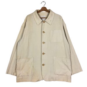Può includere: Giacca beige con colletto, quattro bottoni e due ampie tasche frontali. La giacca è appesa a una gruccia di legno. Il tessuto ha una sottile trama verticale.