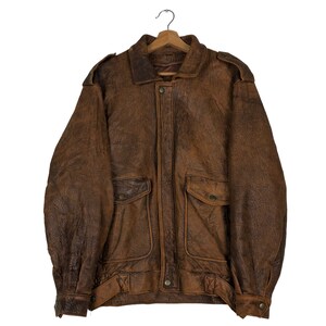 May include: A brown leather jacket with a textured finish, featuring a collared neckline, two front pockets with button closures, and shoulder epaulettes. The jacket is hanging on a wooden hanger.