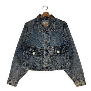 May include: A vintage acid wash denim jacket with a classic collar, two chest pockets, and button closures. The jacket is hung on a wooden hanger. The denim has a blue and white marbled effect.