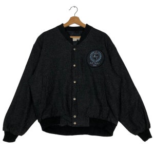 May include: A dark gray bomber jacket with a textured pattern, featuring a black collar, cuffs, and hem. The jacket has a circular patch on the chest with the text "ASTRAY 1960" and a design. It is buttoned up and hanging on a wooden hanger.