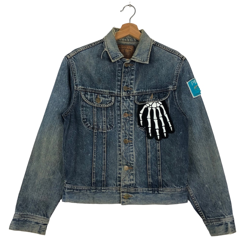 Puede incluir: Una chaqueta vaquera azul con un parche de mano de esqueleto en el bolsillo y un parche azul con el texto "PREMIUM" en la manga.