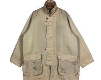 Könnte beinhalten: Eine beige Jacke mit Reißverschluss und mehreren Knopfverschlüssen. Die Jacke hat zwei große Taschen vorne und einen hohen Kragen.