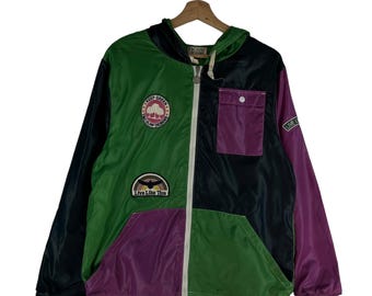 May include: A multi-colored hooded jacket with a zipper closure. The jacket is made of a nylon fabric and has a patchwork design. The jacket has a green, black, and purple color scheme. The jacket has a pocket on the left side and a patch on the right side that says "Keep Green, Declare Designs". The jacket also has a patch on the bottom left side that says "Live Like This".