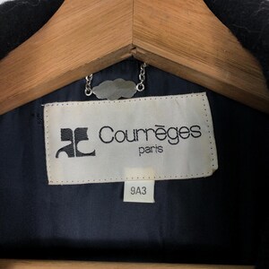 May include: Close-up of a black garment's interior, revealing a label with the text "Courr&egrave;ges paris" in black font. The label is rectangular with a white background and a logo. A silver chain and a wooden hanger are visible.