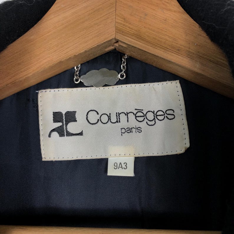 May include: Close-up of a black garment's interior, revealing a label with the text "Courr&egrave;ges paris" in black font. The label is rectangular with a white background and a logo. A silver chain and a wooden hanger are visible.