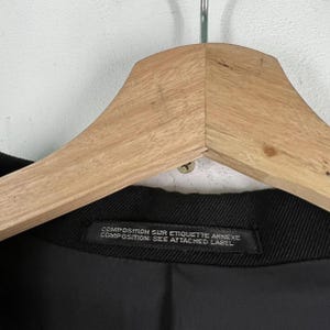 May include: Close-up of a black garment hanging on a wooden hanger. The garment has a label with text that reads "COMPOSITION SUR ETIQUETTE ANNEXE COMPOSITION: SEE ATTACHED LABEL". The background is a light gray wall.
