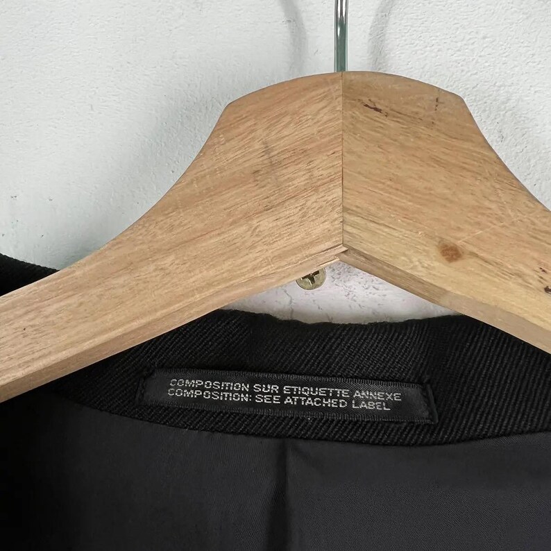 May include: Close-up of a black garment hanging on a wooden hanger. The garment has a label with text that reads "COMPOSITION SUR ETIQUETTE ANNEXE COMPOSITION: SEE ATTACHED LABEL". The background is a light gray wall.