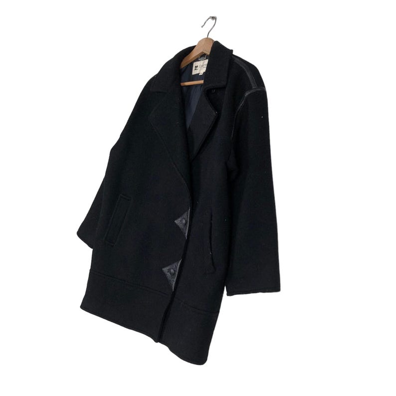 May include: A black wool coat with a double-breasted design, hanging on a wooden hanger. The coat features leather accents on the shoulders and decorative closures. The coat is a classic outerwear piece.