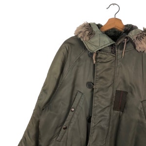 Op de afbeelding: Olijfgroene winterjas met een met bont gevoerde capuchon, knoopsluitingen en een ritssluiting. De jas hangt aan een houten kleerhanger. Een donkerbruin patch is zichtbaar aan de rechter onderkant.