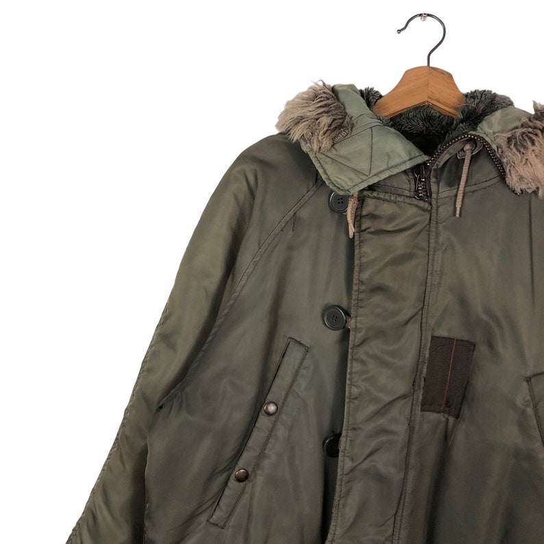 Op de afbeelding: Olijfgroene winterjas met een met bont gevoerde capuchon, knoopsluitingen en een ritssluiting. De jas hangt aan een houten kleerhanger. Een donkerbruin patch is zichtbaar aan de rechter onderkant.