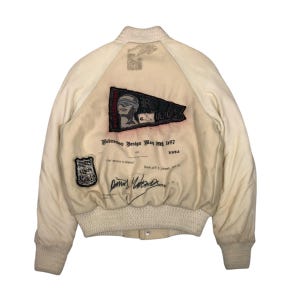 Peut inclure: Veste de bombardier crème avec col, poignets et ourlet côtelés. Le dos présente un écusson avec une figure aux yeux bandés et du texte, ainsi que des écussons supplémentaires et du texte manuscrit. La veste a une fermeture à boutons.