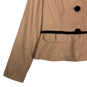 Op de afbeelding: Een beige blazer met zwarte fluwelen afwerking en twee donkerbruine knopen. De blazer heeft een getailleerde pasvorm met &eacute;&eacute;n zak en een klassiek ontwerp. De mouw is zichtbaar aan de linkerkant van de afbeelding.