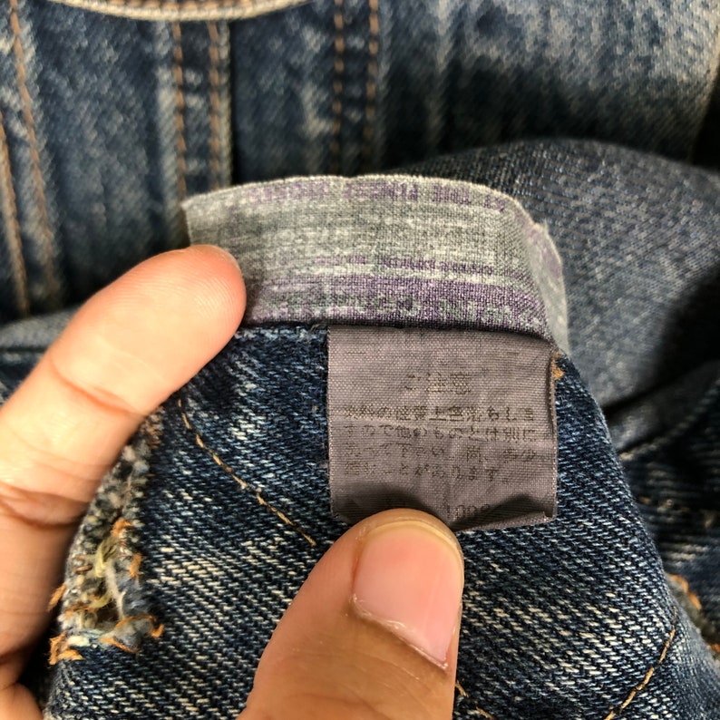 Puede incluir: Primer plano de una etiqueta de mezclilla azul oscuro con texto japon&eacute;s. La etiqueta est&aacute; unida a un par de jeans.